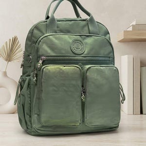 Könnte beinhalten: Ein salbeigrüner Rucksack aus Nylon mit zwei großen Vordertaschen, einem Hauptfach mit Reißverschluss und zwei Tragegriffen an der Oberseite. Der Rucksack verfügt über Seitentaschen mit Kordelzug und ein rundes Logo-Emblem auf der Vorderseite.