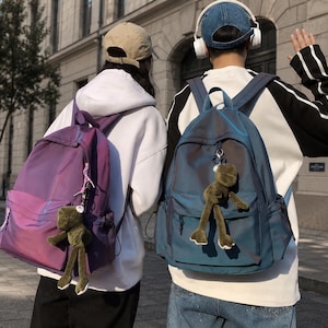 May include: Two backpacks with a shimmering, iridescent finish. One is purple and the other is blue. Each bag features a main zippered compartment, a front pocket, side pockets, and a plush green frog keychain attached to the front zipper pull.