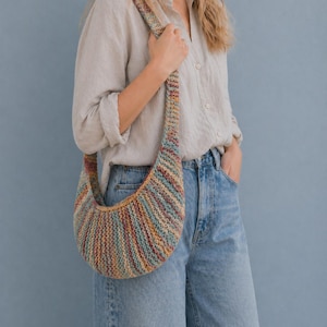 May include: A multicolored, crescent-shaped crochet bag with a long strap. The bag features a striped pattern in shades of blue, red, yellow, and brown. The strap is also striped. The bag is held over the shoulder.