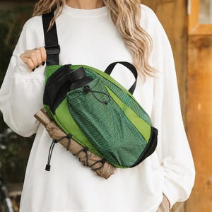 May include: A green crossbody sling bag featuring a black adjustable strap, a black mesh side pocket, and a front drawstring compartment. A black water bottle is tucked into the pocket, and a tan umbrella is secured to the bottom with cords.