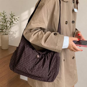 Könnte beinhalten: Eine braune karierte Umhängetasche mit Kordelzug und langem Riemen. Die Tasche besteht aus einem weichen, leichten Material und hat ein großes Fassungsvermögen. Die Tasche wird von einer Person getragen, die einen beigefarbenen Trenchcoat trägt.
