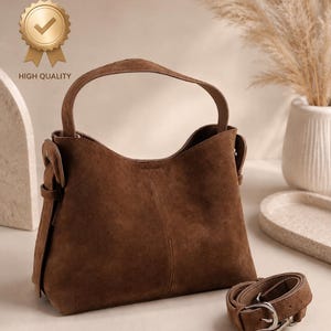 May include: A brown suede handbag with a top handle and an adjustable shoulder strap. The bag has a curved shape and a silver buckle on the strap. The words "HIGH QUALITY" are printed on a gold badge.