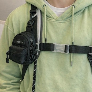 May include: Black crossbody bag with a small front pocket and a zipper closure. The bag is attached to a black strap that goes around the chest. The bag has the text "FASHION & ESS" on it. The person is wearing a light green hoodie.