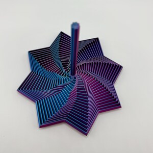 May include: A multi-layered, star-shaped object with a central vertical post. The object features a gradient of colors, transitioning from blue to purple. The design is geometric, with numerous parallel lines creating a layered effect.