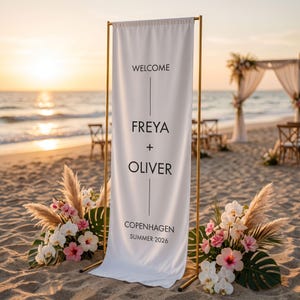 May include: A white welcome sign with gold frame, featuring the names Freya and Oliver, set on a sandy beach. The sign also includes the text "Copenhagen Summer 2026". Floral arrangements with pink and white flowers flank the sign.