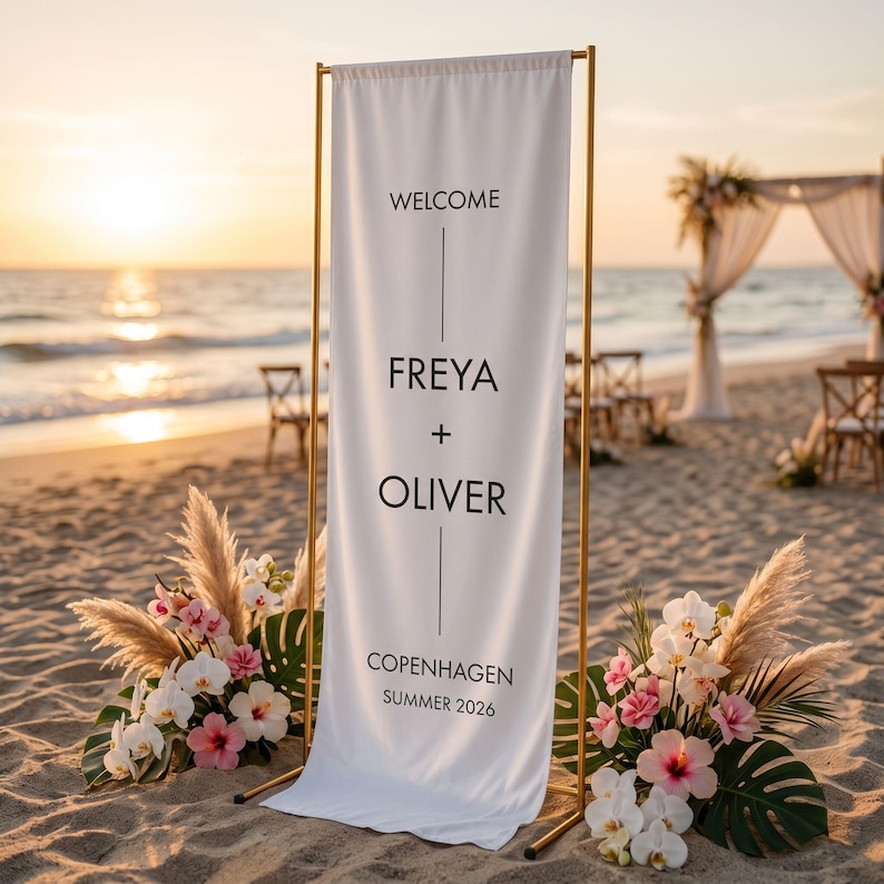 May include: A white welcome sign with gold frame, featuring the names Freya and Oliver, set on a sandy beach. The sign also includes the text "Copenhagen Summer 2026". Floral arrangements with pink and white flowers flank the sign.