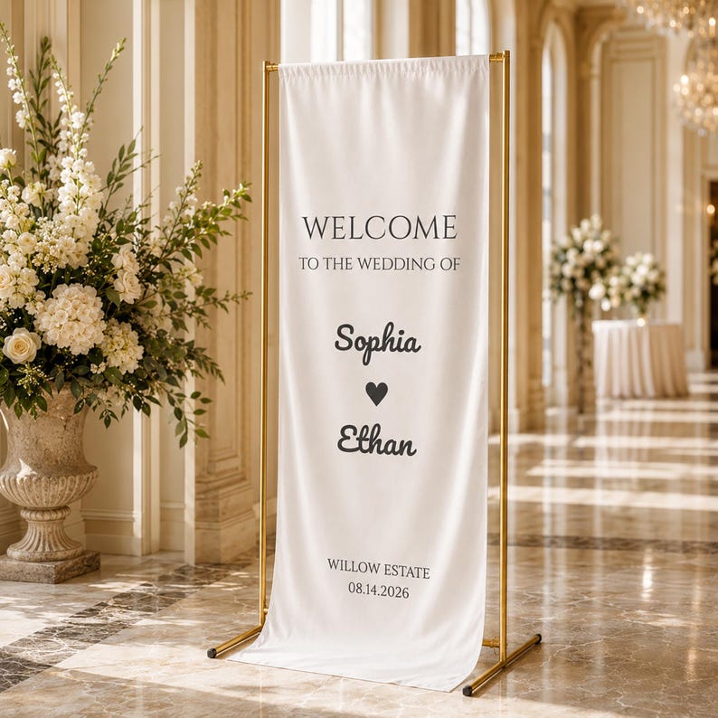 May include: A white wedding welcome sign with gold metal frame. The sign reads "Welcome to the wedding of Sophia & Ethan" with a heart symbol between the names. The date 08.14.2026 and "Willow Estate" are at the bottom.