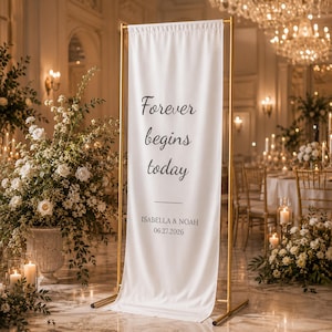 May include: A white banner with gold metal frame, featuring the text "Forever begins today" in black script. Below the text are the names Isabella & Noah and the date 06.27.2026. The banner is displayed in a decorated event space.