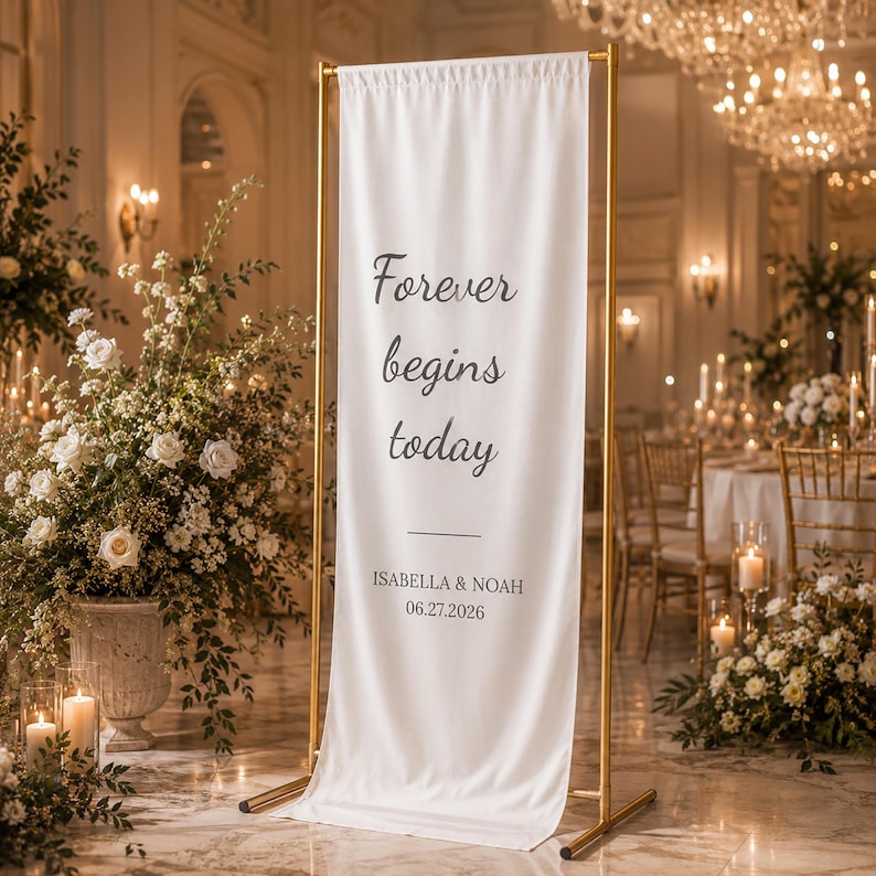 May include: A white banner with gold metal frame, featuring the text "Forever begins today" in black script. Below the text are the names Isabella & Noah and the date 06.27.2026. The banner is displayed in a decorated event space.