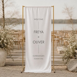 May include: A white fabric welcome sign with gold metal frame. The sign reads "WELCOME" above "FREYA + OLIVER" and "COPENHAGEN SUMMER 2026" in black text. The sign is displayed outdoors near floral arrangements.
