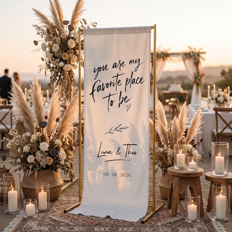 May include: A wedding ceremony sign with the text "you are my favorite place to be" in black script. The sign is on a gold metal frame, with the names "Luna & Theo" and the date 08.08.2026. The scene includes floral arrangements and candles.