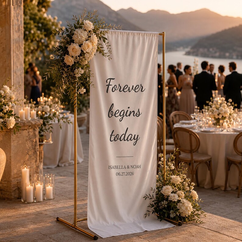 May include: A white banner with the text "Forever begins today" in black script, displayed on a gold metal stand. The banner is decorated with floral arrangements. The names "Isabella & Noah" and the date "06.27.2026" are below the text. The scene is set at an outdoor wedding.
