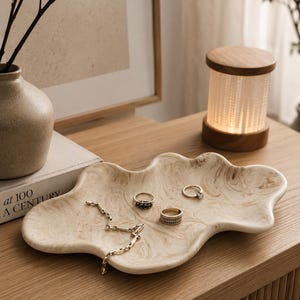 Könnte beinhalten: Ein unregelmäßiges, wolkenförmiges Tablett mit einem beigen und cremefarbenen Marmormuster. Auf dem Tablett liegen eine zarte goldfarbene Kette und drei Ringe, darunter einer mit einem dunklen Stein, einer mit strukturiertem Band und einer mit einem kleinen klaren Stein.