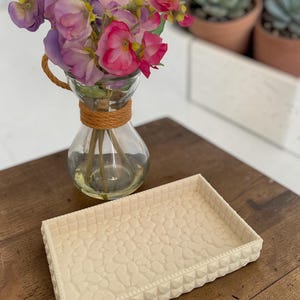 May include: A decorative cream-colored rectangular tray with a textured pebble design and a geometric border. A glass vase with a rope accent holds pink and purple flowers. The items are on a wooden surface.