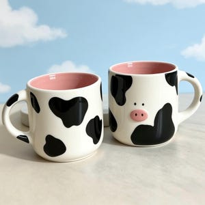 Puede incluir: Dos tazas de cerámica con base blanca y manchas negras de vaca. El interior de cada taza es de color rosa claro. Una taza presenta un pequeño detalle de hocico de cerdo rosa en relieve en la parte frontal, la otra tiene un diseño liso.