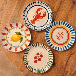 May include: Four decorative ceramic plates with colorful designs. One plate features a red lobster, another a yellow lemon, a third has the text "La dolce vita", and the last one has red tomatoes. Each plate has a unique border.