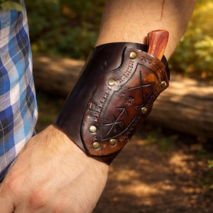Puede incluir: Un brazalete de cuero marrón oscuro con un diseño decorativo y tachuelas de latón, que sostiene un cuchillo de madera. El brazalete se lleva en el antebrazo, con una camisa a cuadros visible en el fondo. Hecho a mano.