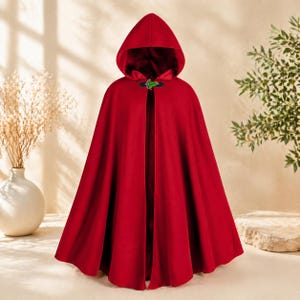 May include: A vibrant red hooded cloak with a matching hood and a decorative clasp. The cloak is made of a soft fabric and drapes elegantly. The clasp is green and black. The cloak is a costume piece.