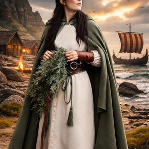 May include: A woman in a white dress and green cloak holds a bundle of greenery. She wears a brown belt and leather arm cuffs. A Viking ship sails on the sea in the background, with a village and mountains in the distance.
