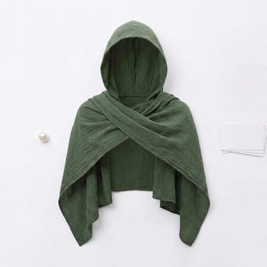Può includere: Un mantello con cappuccio verde oliva scuro realizzato in tessuto testurizzato. Il capo presenta un cappuccio e due ampi pannelli che si incrociano sul petto, creando un aspetto drappeggiato e stratificato con lati aperti e orlo corto.
