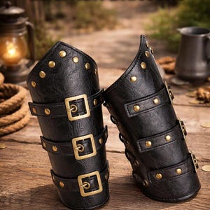 Puede incluir: Brazales de cuero negro con remaches y hebillas doradas. Cada brazal tiene tres correas con hebillas cuadradas. Los brazales se exhiben sobre una superficie de madera. El texto "LEATHER BRACERS" está en la parte superior.