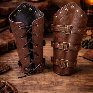 May include: Two brown leather arm cuffs with brass rivets and buckles. One cuff features a lace-up design, while the other has three adjustable straps. The cuffs are designed in a steampunk style.