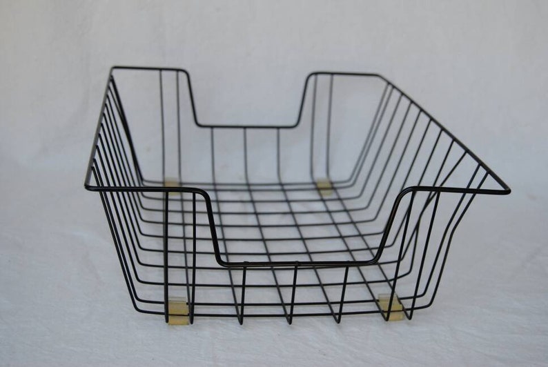 May include: Black wire desk organiser basket with a rectangular shape and a grid pattern. The basket has a curved front and a cutout on one side. The basket is supported by four small, rectangular feet.