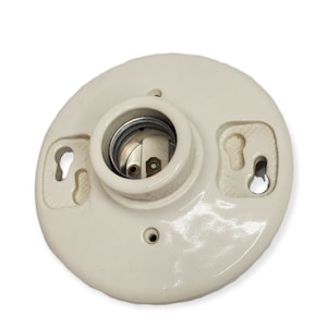 Puede incluir: Base de lámpara de cerámica color crema con un casquillo central y dos ranuras de montaje en forma de ojo de cerradura. La base tiene una forma redondeada con un acabado brillante, diseñada para una fácil instalación. El casquillo metálico es visible en el centro.
