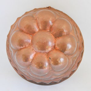 May include: A vintage copper bundt pan with a distinctive, rounded design. The pan features a series of large, bubble-like shapes in the center, surrounded by a decorative edge. The copper has a warm, reddish-brown hue.