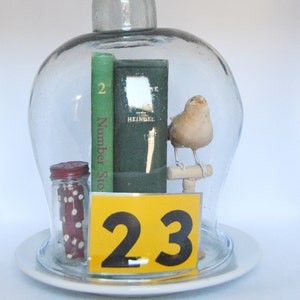 May include: A glass cloche displays a miniature scene. Inside, a small bird perches on a wooden dowel, flanked by two books and a jar of dice. A yellow sign with the number 23 is also present. The cloche sits on a white plate.