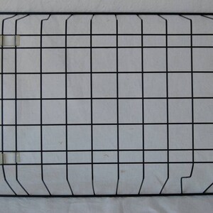 May include: A rectangular black metal grid with a square pattern, secured with clear plastic clips. The grid is designed for wall hanging. The background is a white fabric.