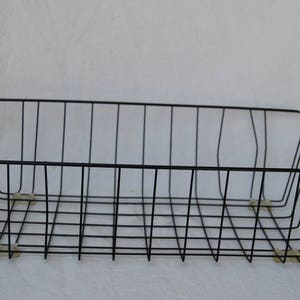 May include: A black wire basket, rectangular in shape with a grid design. The basket has a low profile with slightly raised sides and small feet. The minimalist design is suitable for storage or display, measuring approximately 30 cm in length.