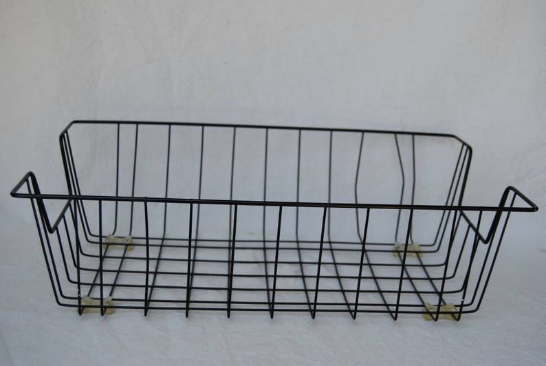 May include: A black wire basket, rectangular in shape with a grid design. The basket has a low profile with slightly raised sides and small feet. The minimalist design is suitable for storage or display, measuring approximately 30 cm in length.