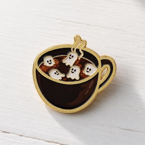 May include: A gold-toned metal enamel pin shaped like a dark brown coffee mug filled with a dark liquid. The mug contains four small, white, smiling ghost-shaped marshmallows floating on the surface, with one ghost rising as steam.