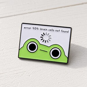 Puede incluir: Un pin de esmalte rectangular con fondo blanco y el texto negro error 404: brain cells not found sobre un icono de carga gris. Debajo, se muestra una cabeza de rana de color verde brillante con dos grandes ojos negros y una boca lineal.