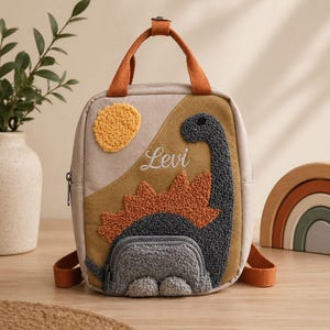 Può includere: Uno zaino beige e marrone con manico e spallacci arancioni. Presenta un design a dinosauro con il nome "Levi" ricamato sul davanti. Il dinosauro è grigio e arancione, con un sole giallo.