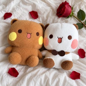 May include: Two plush bear toys, one brown with yellow cheeks and one white with pink cheeks and a brown bow tie, both featuring dark brown eyes and a small mouth with a tongue, surrounded by scattered red rose petals and a single red rose.