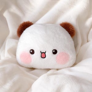 May include: A plush toy shaped like a rounded head with white fur, two brown fuzzy ears, and an embroidered face featuring black eyes, a small mouth with a pink tongue, and two circular pink patches on the cheeks, creating a soft texture.