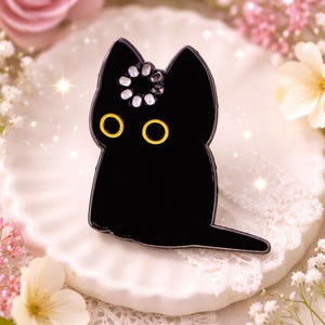 Può includere: Spilla smaltata a forma di gatto nero con occhi gialli e un gruppo decorativo di cristalli sulla testa. La spilla è posta su un piatto bianco smerlato, circondato da fiori.