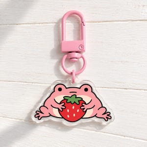 May include: A pink acrylic keychain featuring a cartoon frog holding a red strawberry with a green stem. The frog has a light pink belly, black eyes, and a small mouth. The charm is attached to a pink plastic lobster clasp and a circular ring.