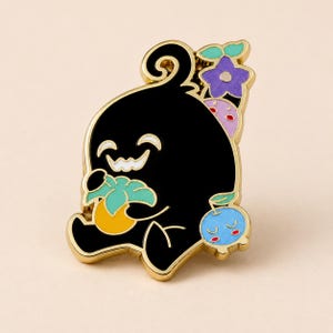 Puede incluir: Un broche de esmalte con borde de metal dorado que muestra una criatura negra con una amplia sonrisa dentada. La criatura sostiene una fruta amarilla con hojas verdes. Arriba hay una flor morada y a la derecha una pequeña fruta azul con cara.