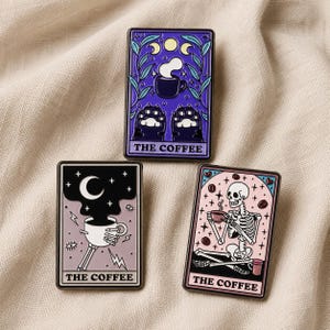 May include: Three rectangular enamel pins featuring tarot card designs with the text THE COFFEE. One pin shows cat paws and a mug, another a skeleton hand holding a mug, and the third a skeleton drinking coffee, all in purple, pink, and black.