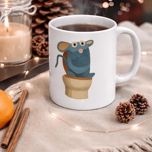 May include: A white ceramic mug with a black handle and the text "But First, Coffee" in a bold, black font. The mug is set against a blurred background, suggesting a cosy setting.