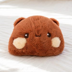 May include: A plush toy shaped like a rounded animal with soft brown fur, two small ears, dark brown embroidered eyes, and two light cream-colored circular patches on the cheeks. The toy has a simple, stitched mouth and a compact, squarish form.