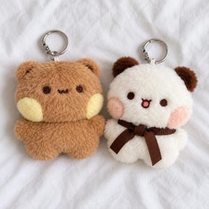 May include: Two plush bear keychains with metal rings. The left bear is brown with yellow cheeks and a simple embroidered face. The right bear is white with brown ears, pink cheeks, a small embroidered mouth, and a brown ribbon bow.