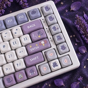 May include: A white keyboard with custom keycaps. The keycaps are a mix of white, lavender, and gray. Some keys feature cat illustrations, paw prints, and the words "BACK", "ENTER", and "SHIFT".