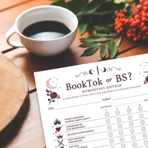 May include: A white ceramic mug filled with dark coffee sits next to a printed rating sheet titled "BookTok or BS?" The sheet features a floral design and prompts for rating various book elements. A wooden cutting board and foliage are in the background.