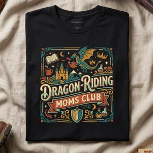 May include: Black t-shirt with a colorful graphic that reads "Dragon Riding Moms Club." The design includes a dragon, castle, books, and potion bottles. The shirt has a crew neck and is made of a soft material.