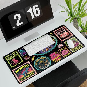 Op de afbeelding: Een rechthoekige zwarte bureaumat met een kleurrijke collage van postzegelachtige illustraties. De ontwerpen bevatten een dinosaurus met de tekst MONGO IS APPALLED, een kat met het label BEST IN DUNGEON en een gekroonde kat.