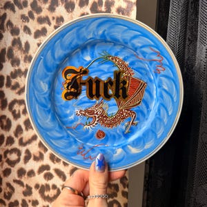 May include: A round ceramic plate with a blue swirled background featuring a brown dragon illustration and the word Fuck in black gothic lettering. The plate has a thin gold rim and a decorative design that covers the centre of the surface.